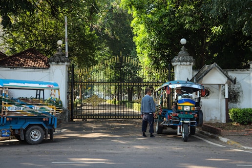 A man stands near a blue and red tuk-tuk parked in front of an ornate, closed iron gate. The area is surrounded by lush green trees and there is another tuk-tuk parked nearby. The architecture has a historic feel, and sunlight filters through the trees, casting dappled shadows on the ground.