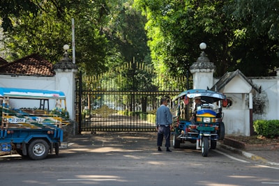 A man stands near a blue and red tuk-tuk parked in front of an ornate, closed iron gate. The area is surrounded by lush green trees and there is another tuk-tuk parked nearby. The architecture has a historic feel, and sunlight filters through the trees, casting dappled shadows on the ground.