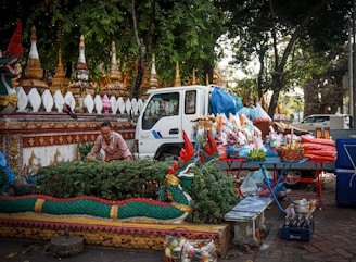 A street vendor is arranging items on a table beside a small white truck. The table is filled with colorful offerings, including candles and bags of items, possibly for a religious or cultural ceremony. In the background, traditional temple structures with ornate designs and vivid colors are visible, surrounded by lush green trees. A decorative dragon-like sculpture frames the scene in the foreground.