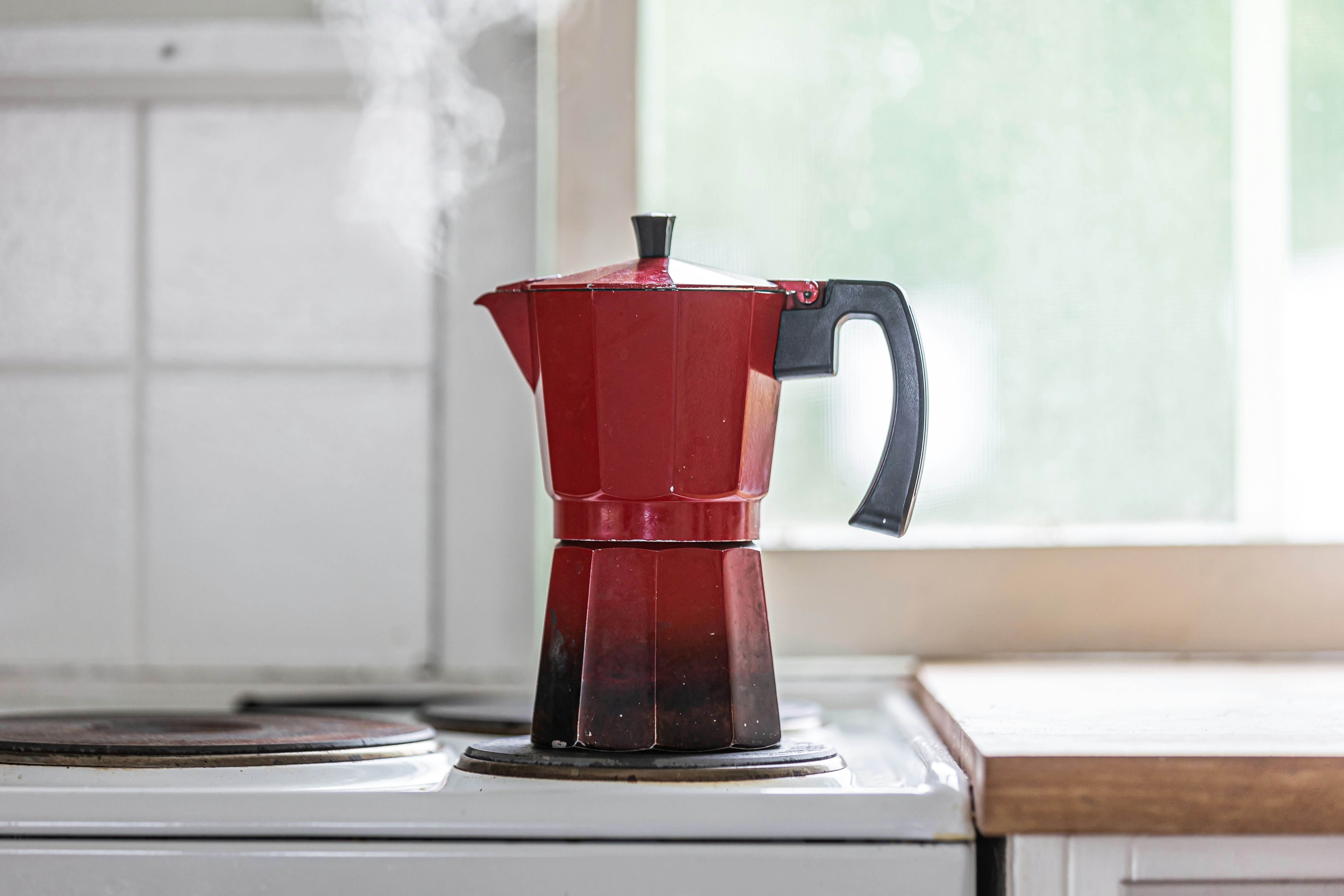 Red moka pot on a stovetop releasing steam while brewing coffee