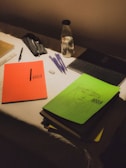 A dimly lit desk contains an assortment of study materials. An orange notebook titled 'Urban Edge' is placed next to a green notebook with a sketched face. Several pens, an eraser, and a ruler are scattered across the surface. A pencil case and a bottle with a black lid labeled 'Frozen' are also present.