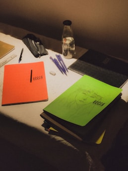 A dimly lit desk contains an assortment of study materials. An orange notebook titled 'Urban Edge' is placed next to a green notebook with a sketched face. Several pens, an eraser, and a ruler are scattered across the surface. A pencil case and a bottle with a black lid labeled 'Frozen' are also present.