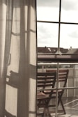 Curtains softly filter the light coming through a large window, with a clear view of an outdoor area featuring wooden chairs arranged on a balcony. The atmosphere suggests a peaceful and reflective moment, with shadows cast on the fabric adding depth.