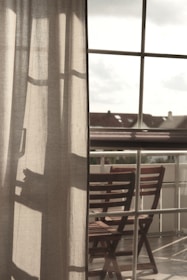 Curtains softly filter the light coming through a large window, with a clear view of an outdoor area featuring wooden chairs arranged on a balcony. The atmosphere suggests a peaceful and reflective moment, with shadows cast on the fabric adding depth.