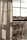 Curtains softly filter the light coming through a large window, with a clear view of an outdoor area featuring wooden chairs arranged on a balcony. The atmosphere suggests a peaceful and reflective moment, with shadows cast on the fabric adding depth.