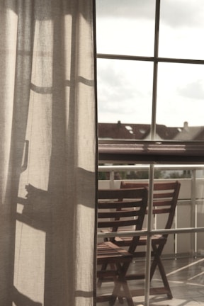 Curtains softly filter the light coming through a large window, with a clear view of an outdoor area featuring wooden chairs arranged on a balcony. The atmosphere suggests a peaceful and reflective moment, with shadows cast on the fabric adding depth.