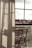 Curtains softly filter the light coming through a large window, with a clear view of an outdoor area featuring wooden chairs arranged on a balcony. The atmosphere suggests a peaceful and reflective moment, with shadows cast on the fabric adding depth.