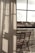 Curtains softly filter the light coming through a large window, with a clear view of an outdoor area featuring wooden chairs arranged on a balcony. The atmosphere suggests a peaceful and reflective moment, with shadows cast on the fabric adding depth.