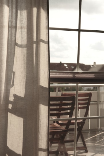 Curtains softly filter the light coming through a large window, with a clear view of an outdoor area featuring wooden chairs arranged on a balcony. The atmosphere suggests a peaceful and reflective moment, with shadows cast on the fabric adding depth.