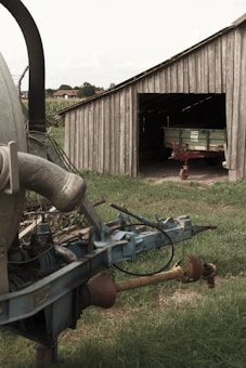 A rustic wooden barn with an open entrance reveals a green and red agricultural trailer inside. To the left, part of a large, weathered piece of farming equipment is visible, with metallic pipes and components. The ground is covered in green grass, and a small part of a rural landscape with houses is visible in the background.