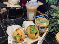 A meal displayed on a shiny black table in a restaurant setting, featuring a banh mi sandwich filled with meat and fresh cilantro, a bowl of Vietnamese noodle soup with meatballs, herbs, and clear broth, and a tall iced coffee in a plastic cup. The items are wrapped in branded packaging with visible text. A leafy green plant is situated near the food, providing a contrast to the dark table surface.