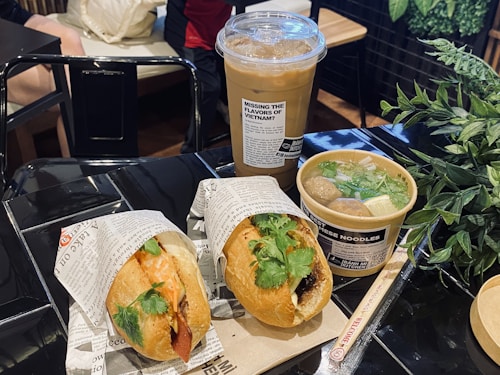 A meal displayed on a shiny black table in a restaurant setting, featuring a banh mi sandwich filled with meat and fresh cilantro, a bowl of Vietnamese noodle soup with meatballs, herbs, and clear broth, and a tall iced coffee in a plastic cup. The items are wrapped in branded packaging with visible text. A leafy green plant is situated near the food, providing a contrast to the dark table surface.