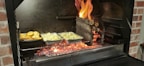 A barbecue grill with glowing coals and flames where logs are burning. Two foil containers are placed on the grill, one containing roasted potatoes and the other cut vegetables. Flames are visible on the right side, creating a vibrant cooking atmosphere.