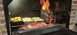A barbecue grill with glowing coals and flames where logs are burning. Two foil containers are placed on the grill, one containing roasted potatoes and the other cut vegetables. Flames are visible on the right side, creating a vibrant cooking atmosphere.