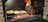 A barbecue grill with glowing coals and flames where logs are burning. Two foil containers are placed on the grill, one containing roasted potatoes and the other cut vegetables. Flames are visible on the right side, creating a vibrant cooking atmosphere.