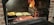 A barbecue grill with glowing coals and flames where logs are burning. Two foil containers are placed on the grill, one containing roasted potatoes and the other cut vegetables. Flames are visible on the right side, creating a vibrant cooking atmosphere.