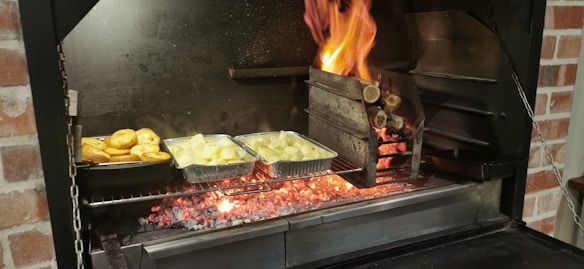 A barbecue grill with glowing coals and flames where logs are burning. Two foil containers are placed on the grill, one containing roasted potatoes and the other cut vegetables. Flames are visible on the right side, creating a vibrant cooking atmosphere.