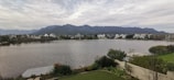 A serene lakeside scene with a row of modern houses in the distance, set against a backdrop of mountains under a cloudy sky. In the foreground, there is a well-maintained garden with a variety of plants and shrubs.