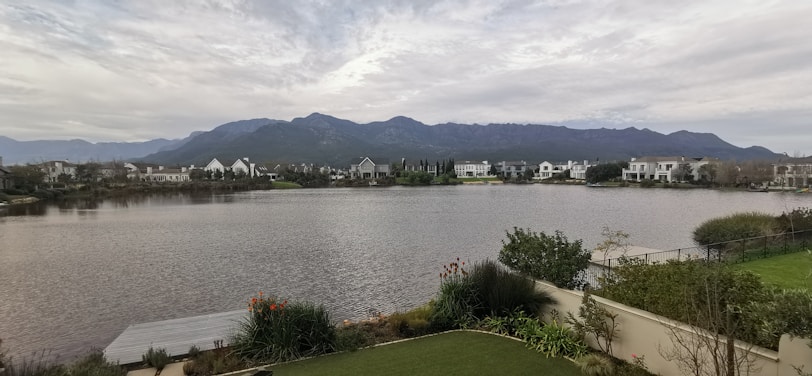 A serene lakeside scene with a row of modern houses in the distance, set against a backdrop of mountains under a cloudy sky. In the foreground, there is a well-maintained garden with a variety of plants and shrubs.
