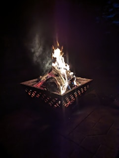 A metal fire pit contains burning logs with flames rising up against a dark background. The fire emits a warm glow that contrasts with the surrounding darkness, creating a cozy atmosphere.