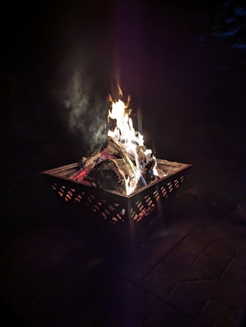 A metal fire pit contains burning logs with flames rising up against a dark background. The fire emits a warm glow that contrasts with the surrounding darkness, creating a cozy atmosphere.