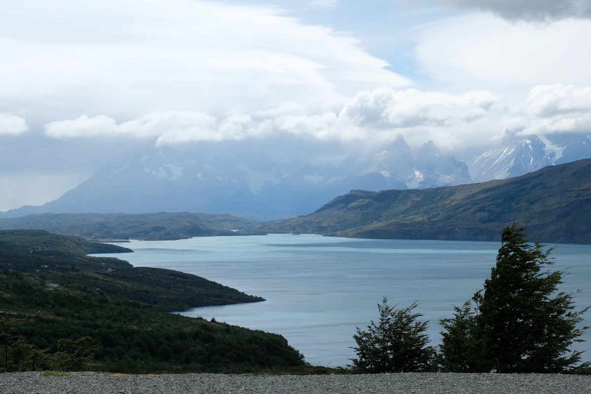 A long blue lake between wooded hills near Torres del Paine, with jagged peaks in cloud behind