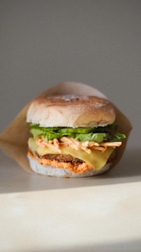 A close-up of a delicious burger with ingredients stacked neatly. The burger includes a toasted bun, crisp lettuce, melted cheese, a creamy sauce, and a meat patty. It is set against a neutral background and positioned slightly off-center, with natural lighting enhancing the textures and colors.