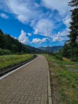 A winding paved pathway runs alongside a set of railroad tracks, surrounded by lush green grass and trees on either side. Tall street lamps line the path, and the sky is a vivid blue filled with fluffy white clouds. Forested hills are visible in the background, creating a sense of tranquility.
