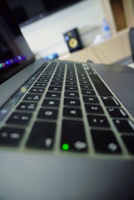 A close-up view of a laptop keyboard, with focus on the keys. The image features a slightly blurred background, showing a monitor with part of an interface visible and a bottle placed alongside.