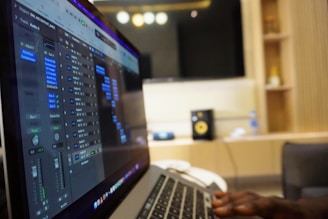 A person is using a laptop with audio editing software open on the screen. The environment suggests a recording or sound studio, with a visible speaker and other equipment in the background shelves. The lighting is warm and subdued.