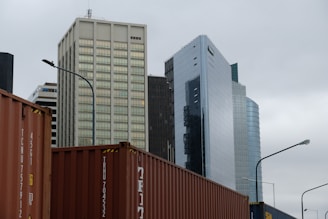 Several modern skyscrapers with varying architectural styles rise against a cloudy sky. In the foreground, multiple shipping containers are visible, hinting at an urban industrial setting. The buildings feature glass and concrete exteriors, reflecting a business district atmosphere.