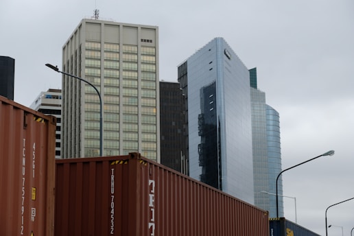 Several modern skyscrapers with varying architectural styles rise against a cloudy sky. In the foreground, multiple shipping containers are visible, hinting at an urban industrial setting. The buildings feature glass and concrete exteriors, reflecting a business district atmosphere.
