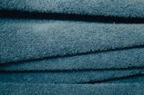 A close-up view of stacked, folded blue towels showing their soft, textured surface. The light catches the fibers, giving the fabric a slightly shiny look.