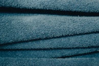 A close-up view of stacked, folded blue towels showing their soft, textured surface. The light catches the fibers, giving the fabric a slightly shiny look.