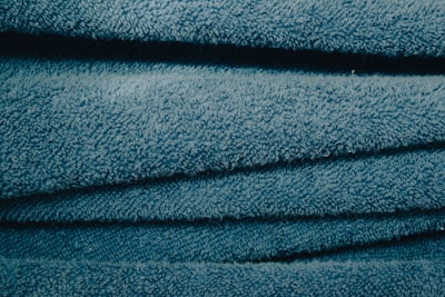 A close-up view of stacked, folded blue towels showing their soft, textured surface. The light catches the fibers, giving the fabric a slightly shiny look.