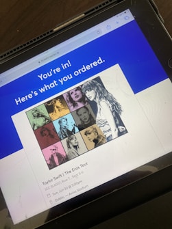 A tablet screen displaying a Ticketmaster confirmation page for a concert ticket purchase. The upper section features a blue banner with white text saying 'You're in! Here's what you ordered.' Below is a collage of colored images showing different portraits of a woman in varying styles and is associated with a concert tour. The event details below mention a tour, location, and time.