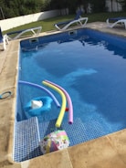A swimming pool with clear water contains several pool toys including colorful pool noodles and an inflatable float. Surrounding the pool are multiple lounge chairs with blue cushions. In the background, there is a grassy area and a white fence. The pool area appears well-maintained and inviting.