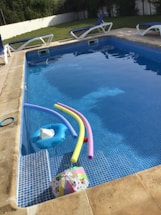 A swimming pool with clear water contains several pool toys including colorful pool noodles and an inflatable float. Surrounding the pool are multiple lounge chairs with blue cushions. In the background, there is a grassy area and a white fence. The pool area appears well-maintained and inviting.