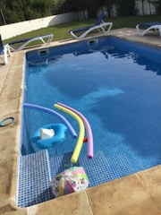 A swimming pool with clear water contains several pool toys including colorful pool noodles and an inflatable float. Surrounding the pool are multiple lounge chairs with blue cushions. In the background, there is a grassy area and a white fence. The pool area appears well-maintained and inviting.