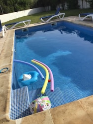 A swimming pool with clear water contains several pool toys including colorful pool noodles and an inflatable float. Surrounding the pool are multiple lounge chairs with blue cushions. In the background, there is a grassy area and a white fence. The pool area appears well-maintained and inviting.