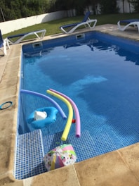 A swimming pool with clear water contains several pool toys including colorful pool noodles and an inflatable float. Surrounding the pool are multiple lounge chairs with blue cushions. In the background, there is a grassy area and a white fence. The pool area appears well-maintained and inviting.