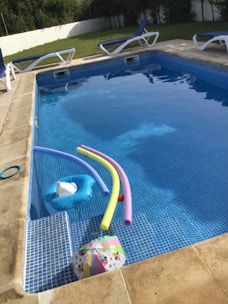 A swimming pool with clear water contains several pool toys including colorful pool noodles and an inflatable float. Surrounding the pool are multiple lounge chairs with blue cushions. In the background, there is a grassy area and a white fence. The pool area appears well-maintained and inviting.