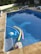 A swimming pool with clear water contains several pool toys including colorful pool noodles and an inflatable float. Surrounding the pool are multiple lounge chairs with blue cushions. In the background, there is a grassy area and a white fence. The pool area appears well-maintained and inviting.