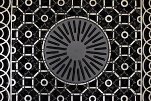 A patterned metal grate features a circular design in the center with radiating slots and a surrounding intricate geometric grid. The grid consists of repeating circular and square shapes connected by intersecting lines.