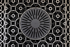 A patterned metal grate features a circular design in the center with radiating slots and a surrounding intricate geometric grid. The grid consists of repeating circular and square shapes connected by intersecting lines.