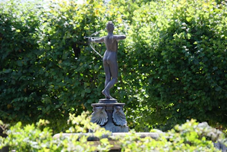 A bronze statue depicting an archer, posed with a bow drawn and aiming. The statue is set against a dense background of lush green foliage, giving the impression of a garden or park setting. Light filters through the leaves, creating dappled shadows on the statue.