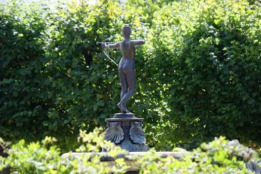 A bronze statue depicting an archer, posed with a bow drawn and aiming. The statue is set against a dense background of lush green foliage, giving the impression of a garden or park setting. Light filters through the leaves, creating dappled shadows on the statue.