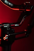A close-up view of a black and red mechanical arm or stabilizer with a label RS3 PRO against a dark red background. The object is illuminated with red lighting, adding a dramatic effect.