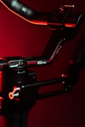 A close-up view of a black and red mechanical arm or stabilizer with a label RS3 PRO against a dark red background. The object is illuminated with red lighting, adding a dramatic effect.