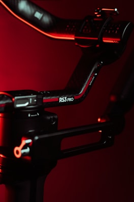 A close-up view of a black and red mechanical arm or stabilizer with a label RS3 PRO against a dark red background. The object is illuminated with red lighting, adding a dramatic effect.